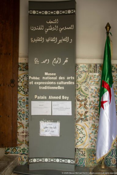Freestanding museum sign beside an Algerian flag and patterned tile walls inside Ahmed Bey Palace.