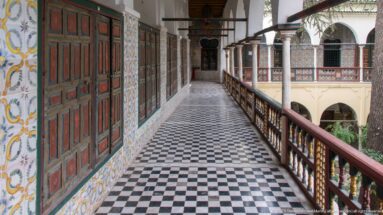 Upper palace gallery with black-and-white checkered floor, painted doors, tiled wall, wooden railing, and courtyard arcade.