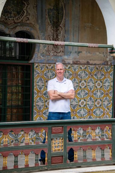 Man in a white polo shirt standing behind a painted balcony railing against patterned tiles and barred windows.