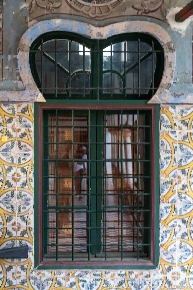 Green window with metal grille and oval barred transom, surrounded by patterned tiles and faded painted wall decoration.
