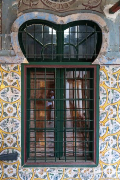 Green window with metal grille and oval barred transom, surrounded by patterned tiles and faded painted wall decoration.