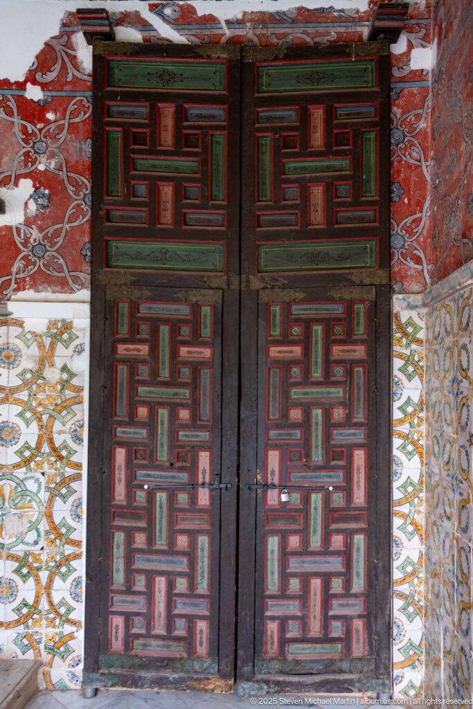 Dark double doors with red and green geometric paneling between patterned tile fields beneath a weathered red painted wall.