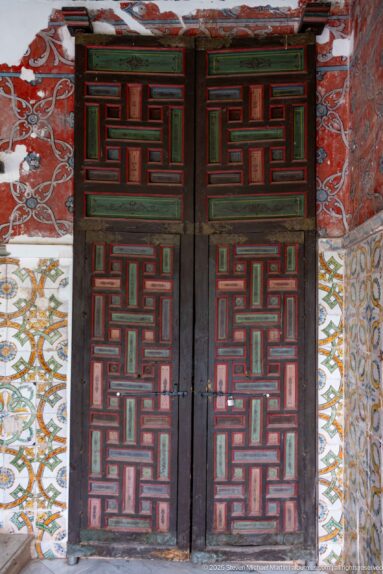 Dark double doors with red and green geometric paneling between patterned tile fields beneath a weathered red painted wall.