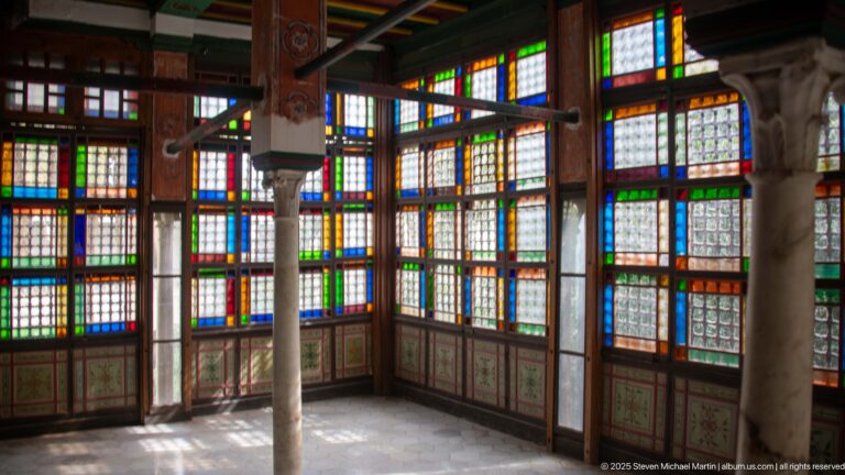 Interior room lined with tall multicolored glass windows, wooden grids, slender columns, and sunlight across the floor.