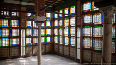 Interior room lined with tall multicolored glass windows, wooden grids, slender columns, and sunlight across the floor.