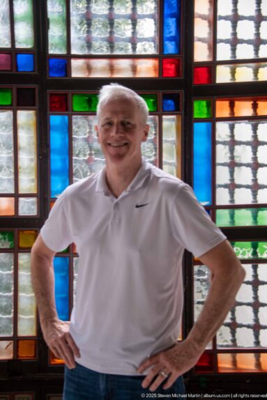 Smiling man in a white polo shirt standing before multicolored red, blue, green, and amber palace windows.