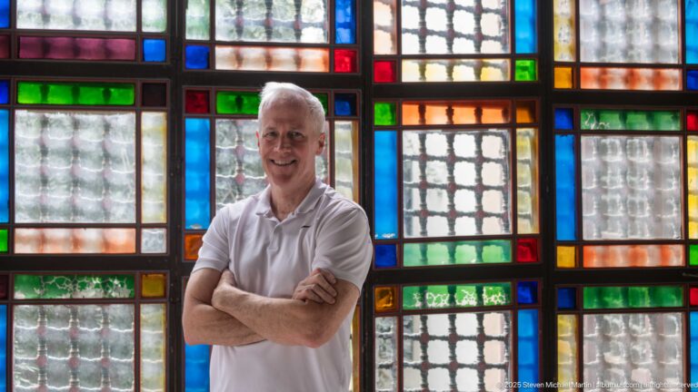 Man with folded arms standing before a close grid of red, blue, green, amber, and textured clear glass panes.