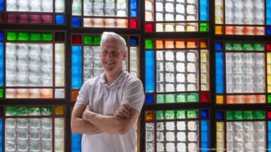 Man with folded arms standing before a close grid of red, blue, green, amber, and textured clear glass panes.