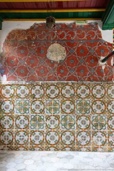 Weathered red painted wall with pale looping motifs above a broad dado of repeating patterned ceramic tiles.