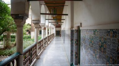 Long palace gallery with patterned tile dado, polished floor, columns, painted ceiling beams, and garden opening at left.