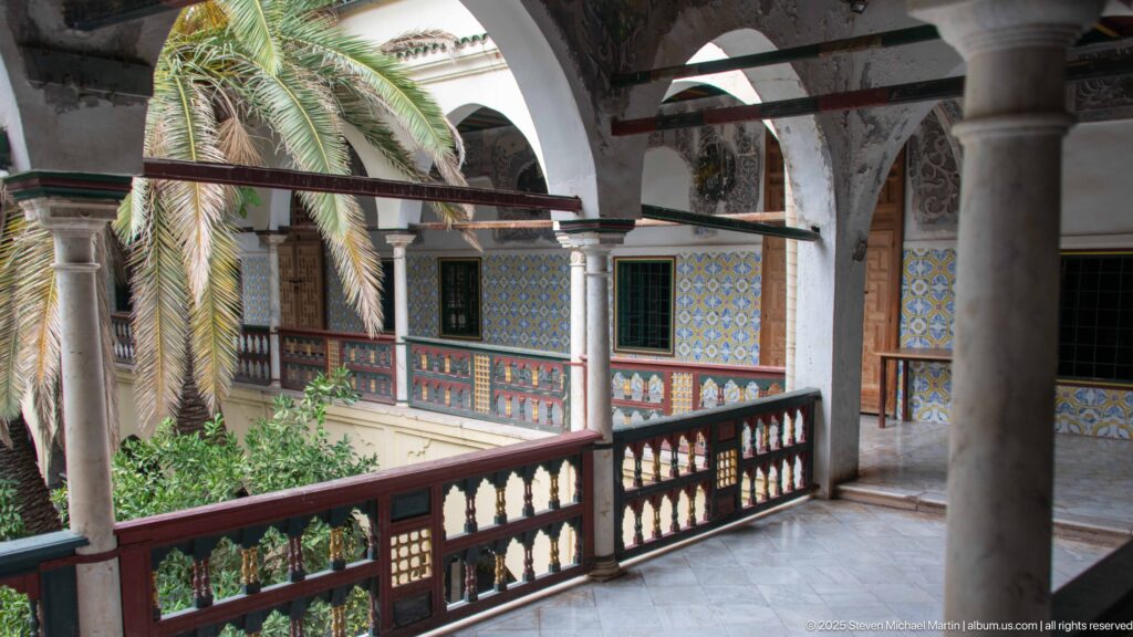Upper palace arcade with stone columns and pointed arches overlooking tiled balconies and palm foliage across the courtyard.