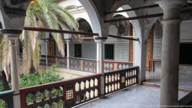 Upper palace arcade with stone columns and pointed arches overlooking tiled balconies and palm foliage across the courtyard.