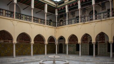 Two-story palace courtyard with pointed arches, tiled lower walls, painted upper railings, slender columns, and central fountain.