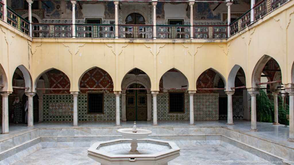 Octagonal courtyard fountain in front of a row of pointed arches, slender columns, tiled walls, and upper gallery.
