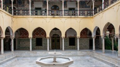 Octagonal courtyard fountain in front of a row of pointed arches, slender columns, tiled walls, and upper gallery.