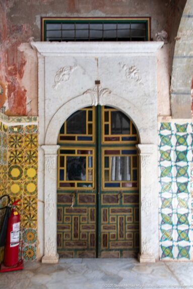 Low geometric double door beneath a pale rounded stone arch, flanked by patterned tiles and a red fire extinguisher.