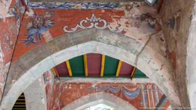 Pointed stone arch beneath weathered red and blue wall paintings, with a second arch and multicolored ceiling beyond.
