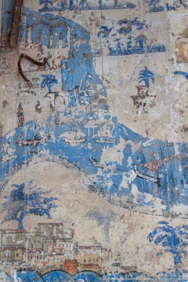 Faded blue wall mural with fragmentary buildings and landscape or maritime forms interrupted by extensive paint loss.