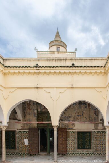 Pale courtyard façade with broad arches, decorated rooms, tiled walls, painted doors, and a small polygonal rooftop pavilion.