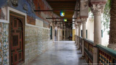 Arcaded palace passage with carved columns, painted wooden door, patterned tile walls, murals, and a palm visible beyond the railing.