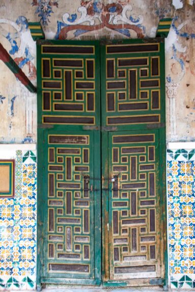 Green double doors with gold geometric bands, surrounded by blue-yellow tiles and weathered wall paintings above.