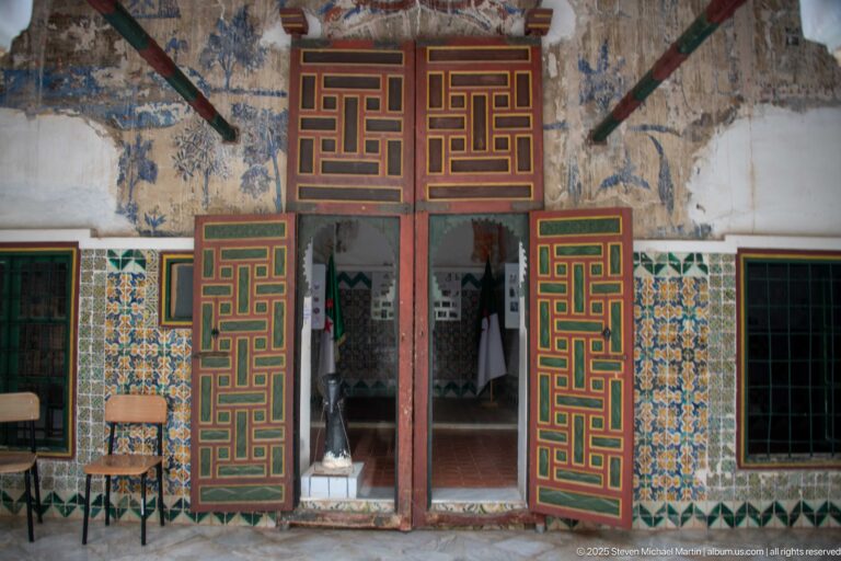 Open red, green, and ochre paneled doors framing a museum room, with patterned tiles and faded murals around the entrance.
