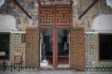 Open red, green, and ochre paneled doors framing a museum room, with patterned tiles and faded murals around the entrance.