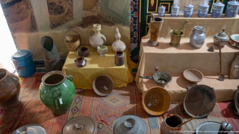 Display of pottery jars, pitchers, cups, metal vessels, sieves, and household utensils arranged on tables and patterned rugs.