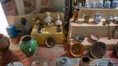 Display of pottery jars, pitchers, cups, metal vessels, sieves, and household utensils arranged on tables and patterned rugs.