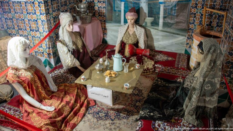 Four female mannequins in traditional dress seated around a low table with serving vessels, cups, carpets, and cushions.