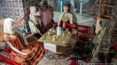 Four female mannequins in traditional dress seated around a low table with serving vessels, cups, carpets, and cushions.