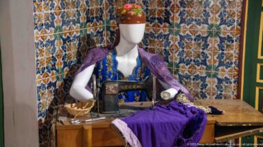 Female mannequin in colorful traditional clothing seated at a vintage sewing machine with purple fabric against patterned tiles.