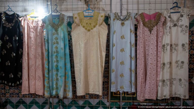Long traditional dresses in cream, blue, pink, black, and white hanging against a tiled wall, many with embroidered necklines.