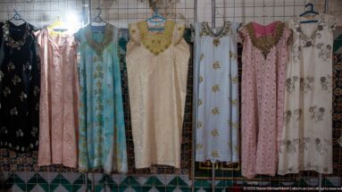 Long traditional dresses in cream, blue, pink, black, and white hanging against a tiled wall, many with embroidered necklines.