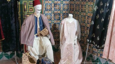 Male and female mannequins in traditional garments standing before patterned tiles, with additional robes hanging at both sides.