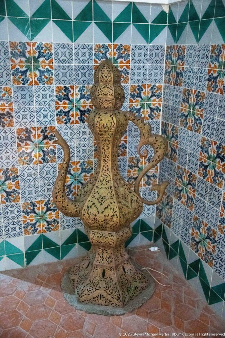 Ornate brass-colored pierced metal vessel with tall neck, looping handles, bulbous body, and flared base against tiled walls.