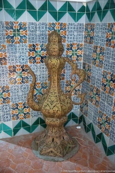 Ornate brass-colored pierced metal vessel with tall neck, looping handles, bulbous body, and flared base against tiled walls.
