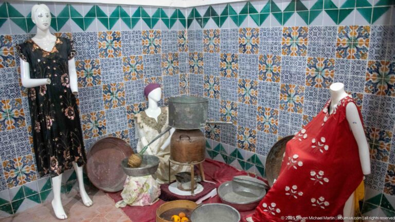 Female mannequins in traditional garments displayed with large trays, pots, bowls, and household vessels against patterned tiles.