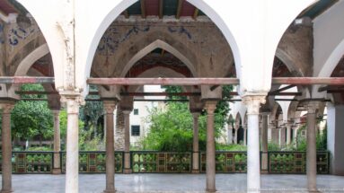 Pointed palace arches on slender stone columns framing a green courtyard and painted balustrade beyond.