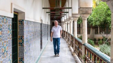 Man walking along a palace gallery with blue-orange tiled wall, stone columns, painted railing, and garden at right.
