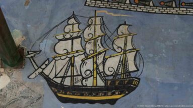 Painted three-masted sailing ship with black rigging, yellow spars, cream sails, and dark hull against blue-gray wall.