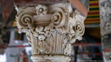 Close-up of carved stone capital with large volutes, curling foliage, drilled details, and weathered edges.