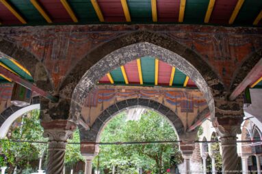Layered pointed arches on carved and spiral-fluted columns beneath a red, green, and yellow ceiling, with garden foliage beyond.