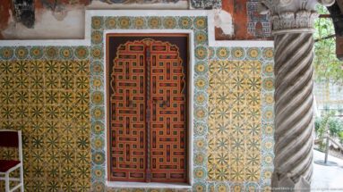 Red geometric double door surrounded by yellow-green patterned tiles beside a large spiral-fluted stone column.