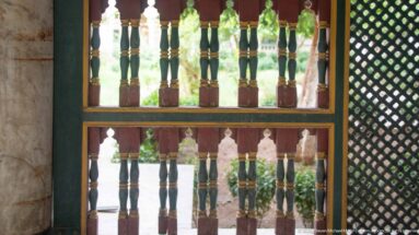 Two rows of multicolored turned wooden balusters beside a dark green lattice screen in a palace gallery.