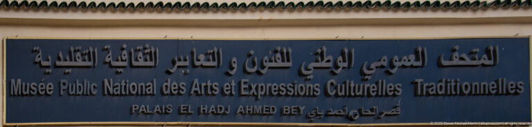 Blue exterior museum sign with Arabic and French lettering naming the National Public Museum and Palais El Hadj Ahmed Bey.