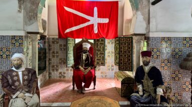 Three male mannequins in traditional dress, one seated beneath a large red banner, against patterned tiles and painted shutters.