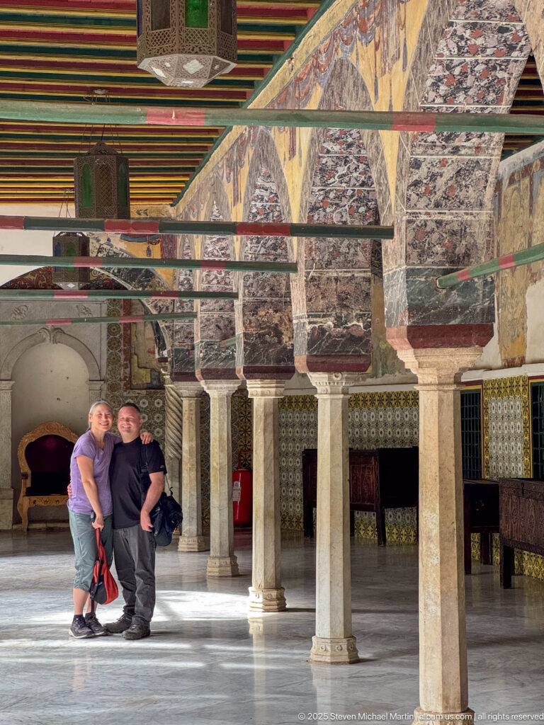 Two men standing beneath painted palace arches with stone columns, patterned tiled walls, and a multicolored ceiling.
