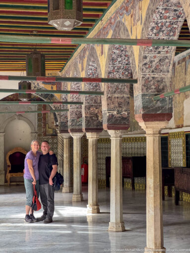 Two men standing beneath painted palace arches with stone columns, patterned tiled walls, and a multicolored ceiling.