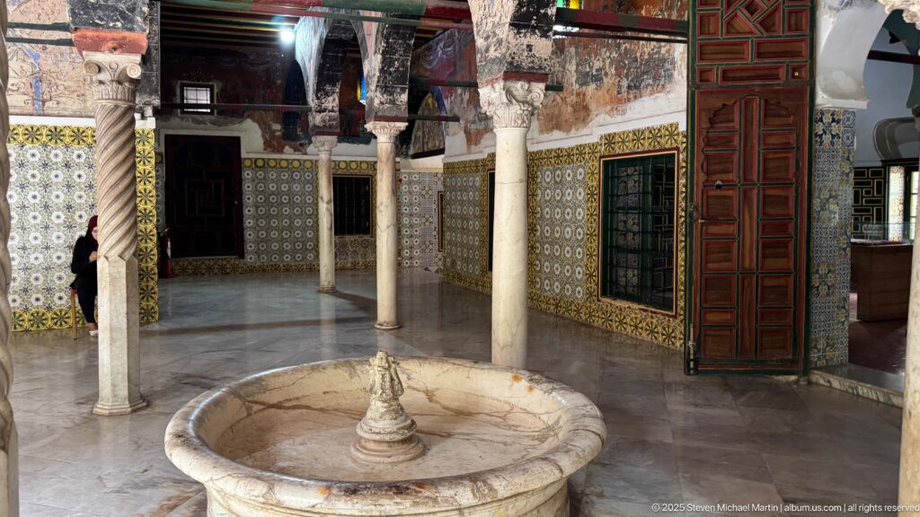 Round stone fountain basin in a palace courtyard surrounded by slender columns, patterned tiles, windows, and a tall wooden door.
